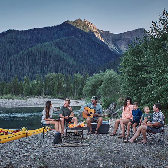 A group of people have a campfire on a rocky beach, with boats docked on the shore.