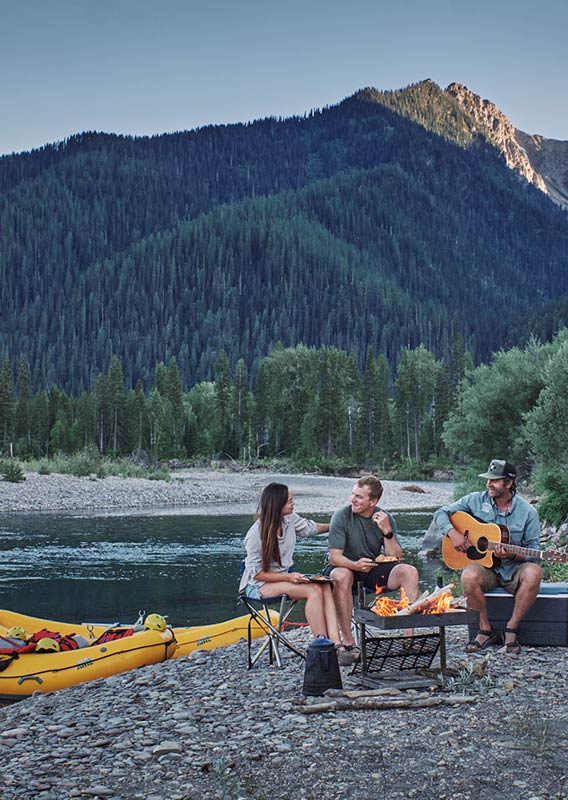 A group of people are camped around a fire on a shoreline, their yellow rafts on the shore.