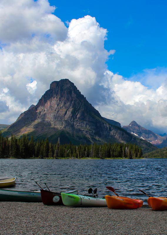 A row of canoes and kayaks on a pebble beach. Mountains across the lake