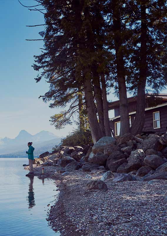 Man canoeing on a lake, woman on shore. Mountains across the lake.