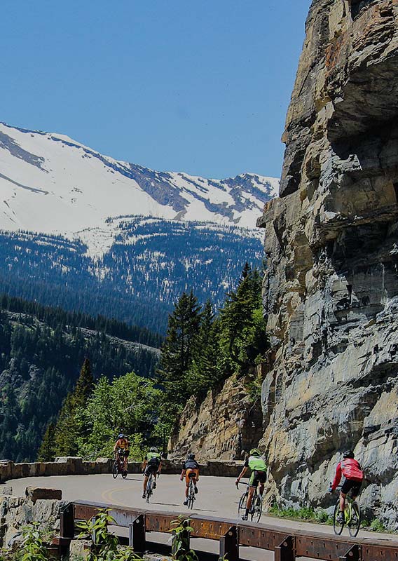 Cyclists ride around a bend in a road under tall rocky cliffs.