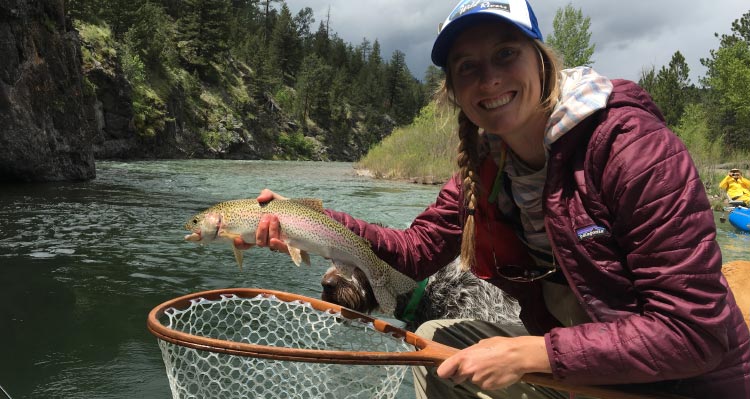 A woman holds a trout and net above a river.