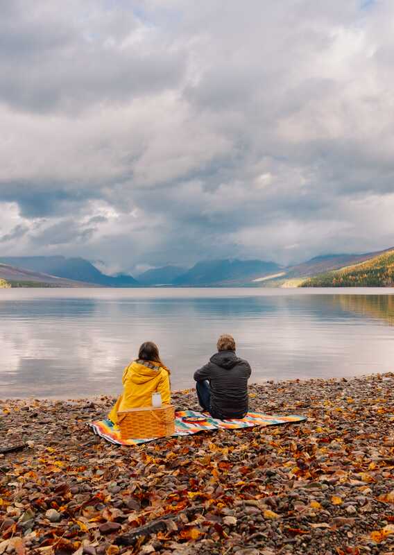 Two people sit on a colourful rocky beach by a large lake.