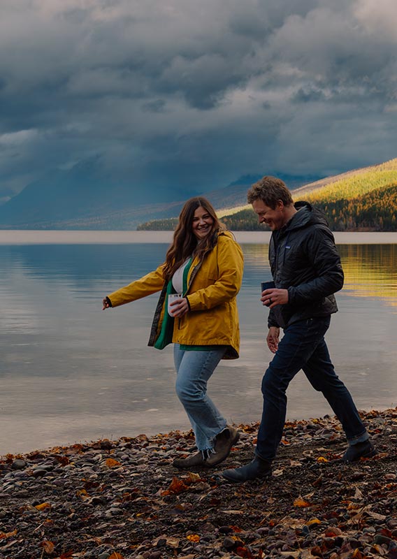Two people walk on a rocky beach next to a large lake.