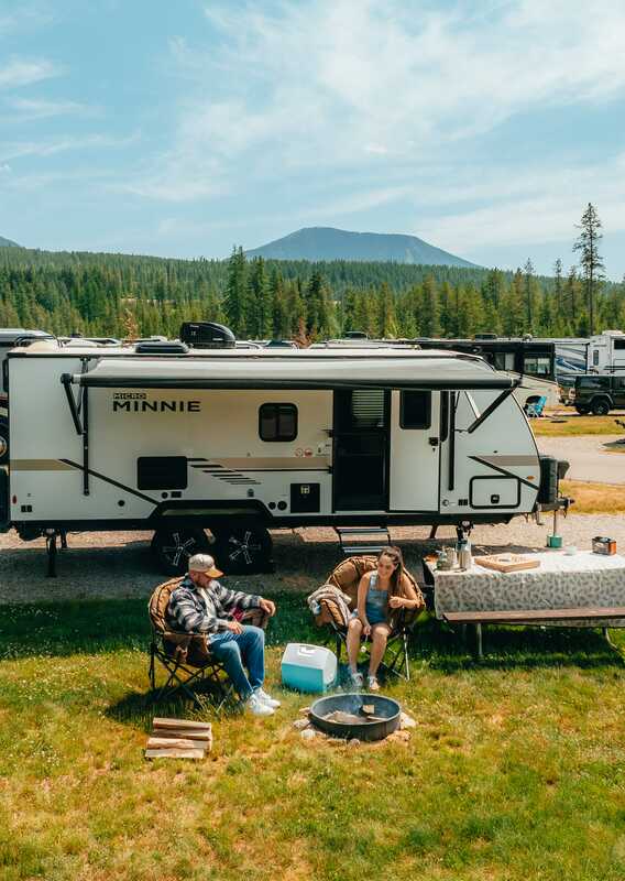 Two people sit outside their RV at a campsite.