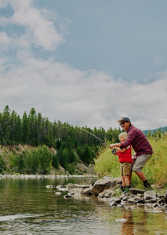 A dad and kid cast a fishing rod from a river's edge.