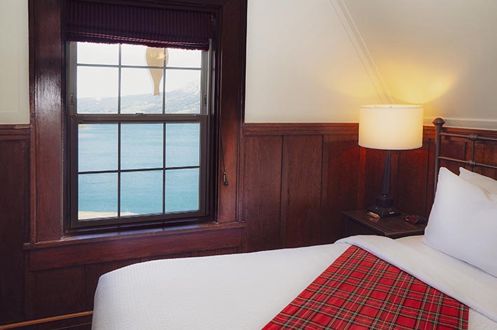 A hotel bed near a window overlooking a blue lake.