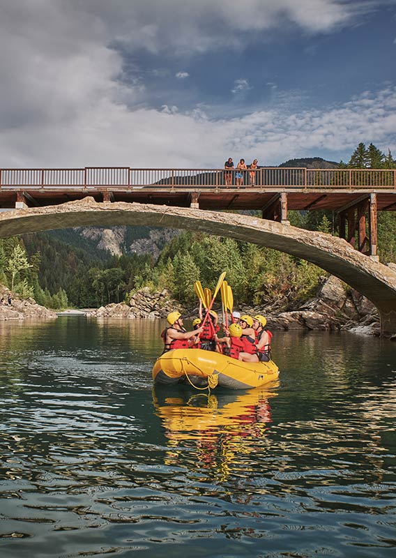 A group of people on a yellow raft on a river passing an old bridge.